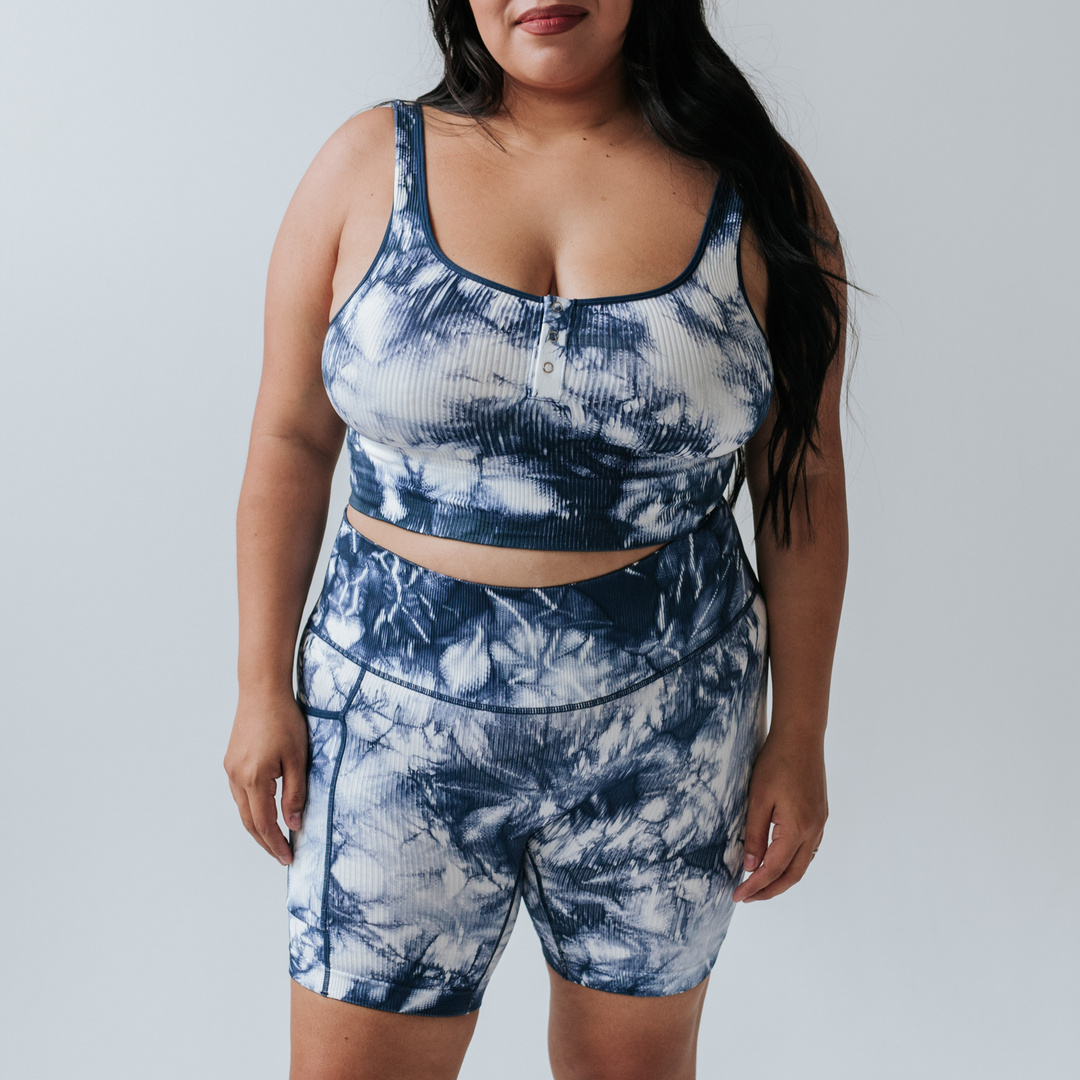 [Night Sky] Plus-size model wearing a ribbed white and blue tie-dye nursing bra with wide straps and a button-front detail for easy access, paired with matching high-waisted bottoms. The bottoms have a pocket on the right hand side. 
